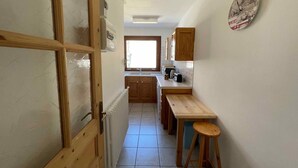 Een smalle keuken met houten keukenkastjes, een radiator, een raam en een wandklok.