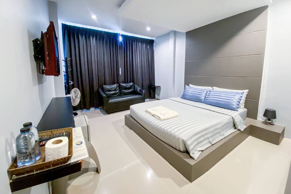 A modern bedroom with a large bed, a black leather sofa, a flat-screen TV mounted on the wall, and a bedside table with a lamp.