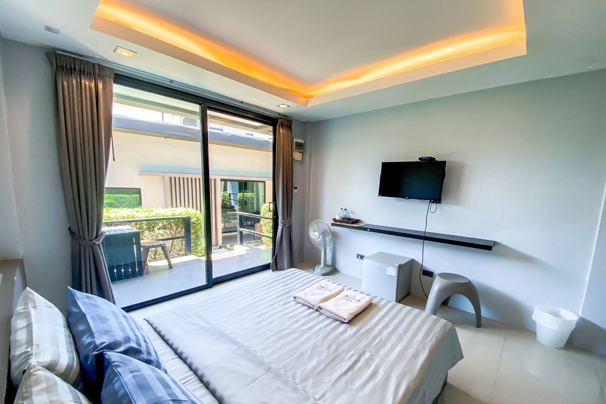 A modern hotel room with a large bed, a flat-screen TV mounted on the wall, a desk with a chair, and a balcony with a view of greenery.