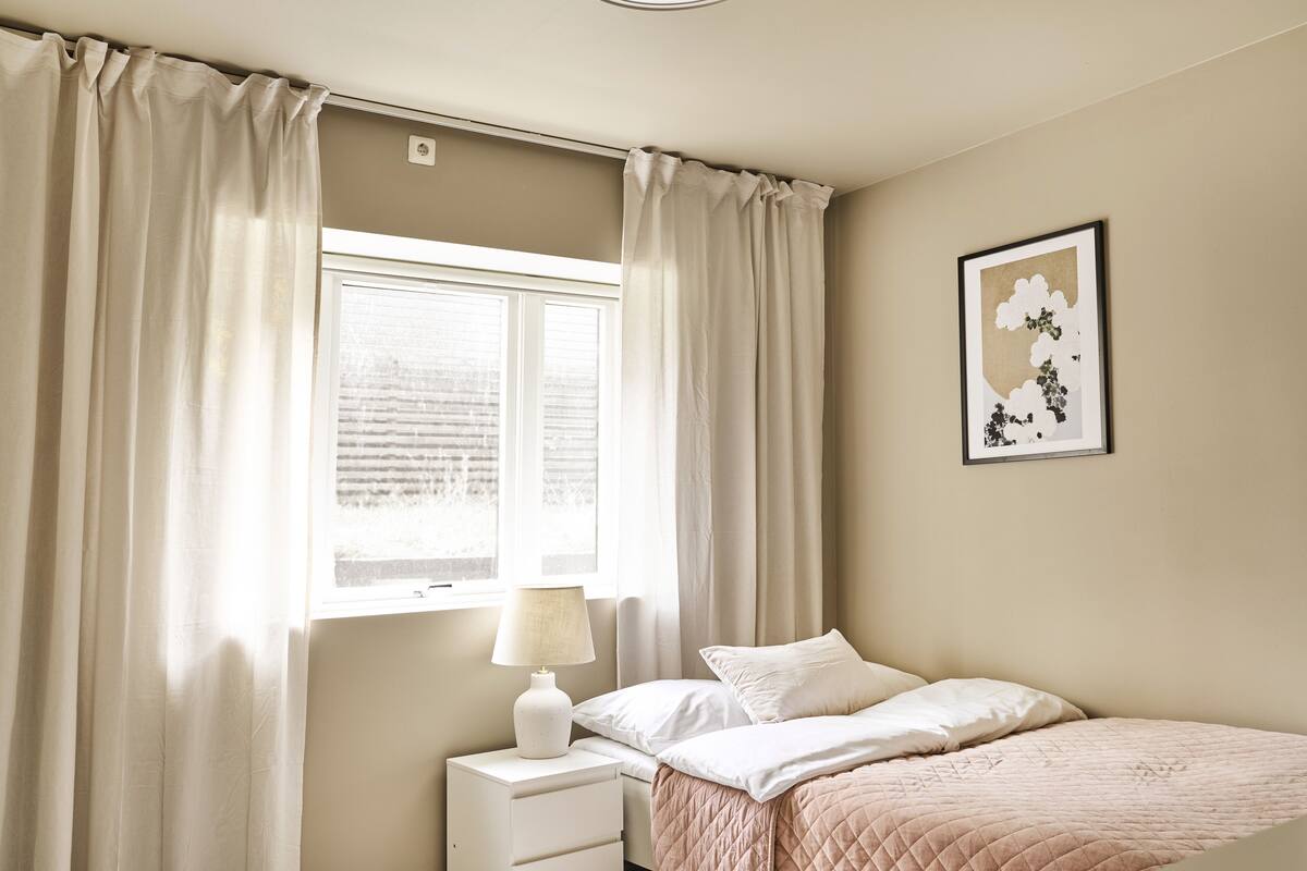 A bedroom with a bed, a nightstand, a lamp, a window with curtains, and a wall-mounted picture.