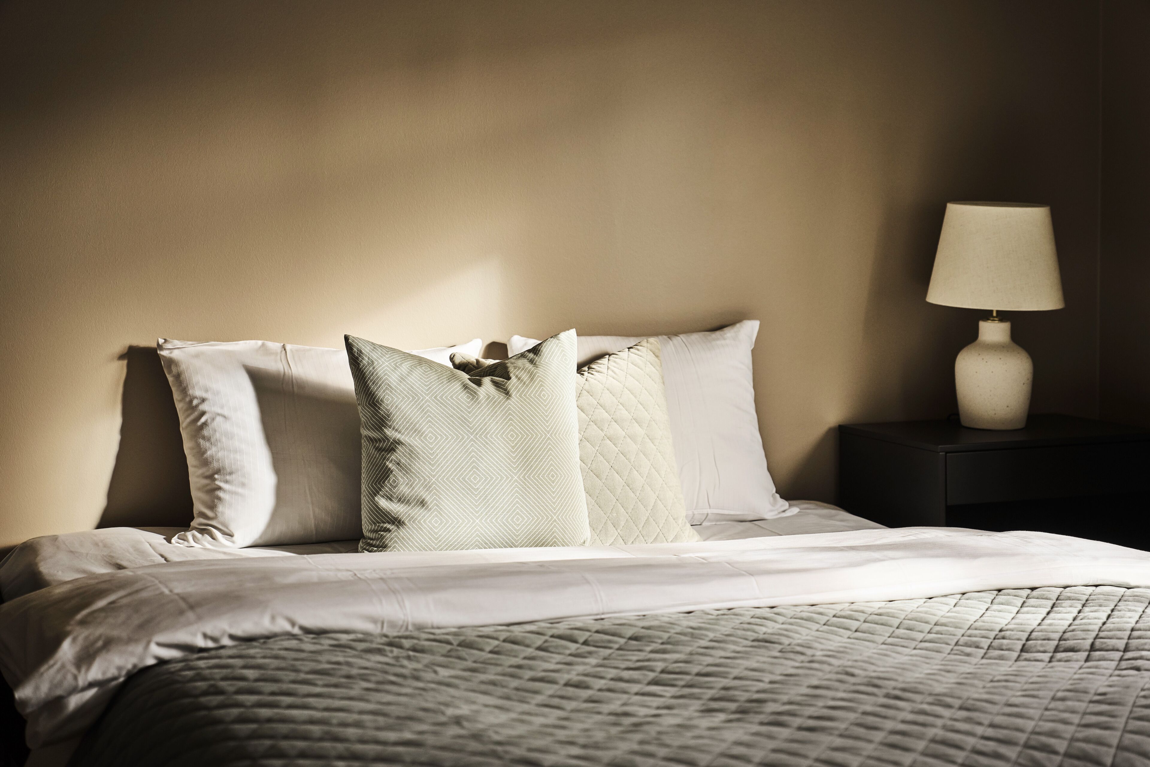 Una cama bien hecha con un edredón acolchado y varias almohadas, una lámpara de noche y una mesita de noche oscura.