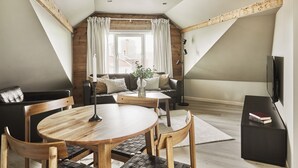 A cozy loft bedroom with a wooden ceiling, a large window with curtains, a black leather sofa, a wooden dining table, and a TV on a black stand. - Staysville – City Center (Stavanger)