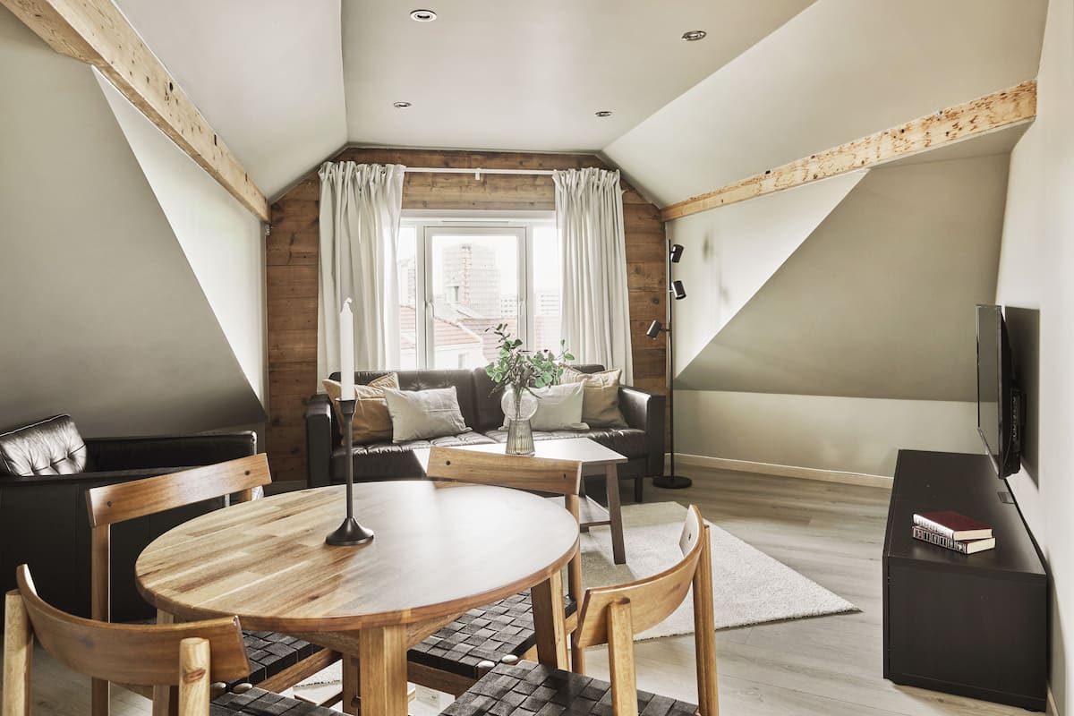 A cozy loft bedroom with a wooden ceiling, a large window with curtains, a black leather sofa, a wooden dining table, and a TV on a black stand.