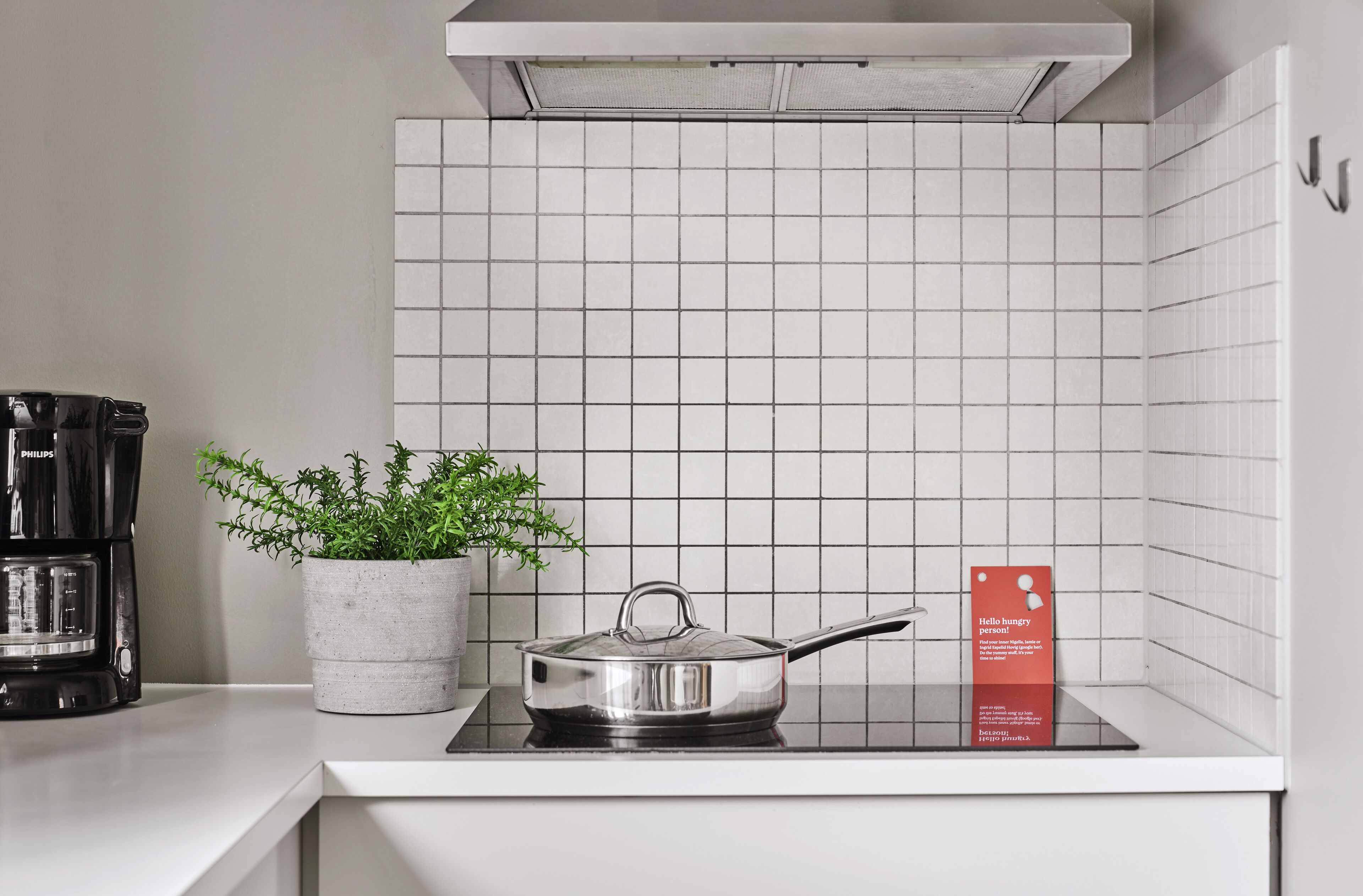 Une cuisine avec un mur recouvert de carreaux blancs, une casserole en acier inoxydable posée sur un plan de travail noir, une plante en pot et un panneau rouge avec du texte.