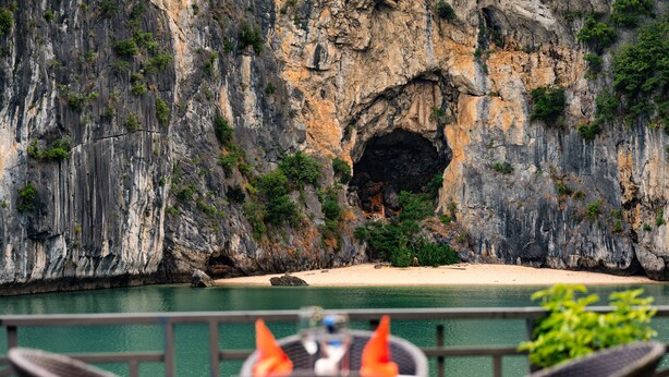 A dining area with wicker chairs and a table, overlooking a coastal cliff with a cave entrance and a sandy beach. - La Pandora Boutique Cruise (Tuan Chau)
