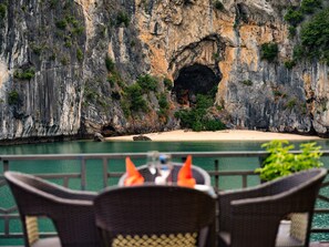 Un comedor con sillas de mimbre y una mesa, con vista a un acantilado costero que incluye la entrada de una cueva y una playa de arena.