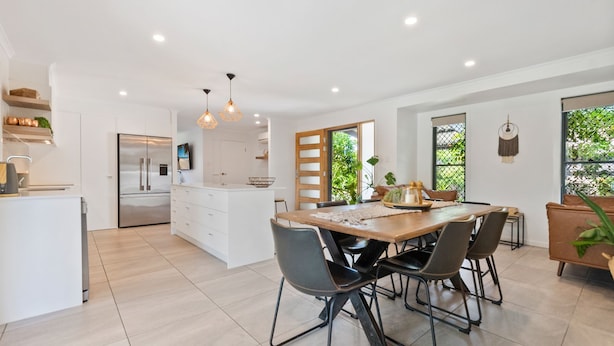 A modern kitchen with a central island, a dining area with chairs, and a large window with a view of greenery.