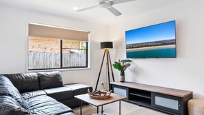 A modern living room with a leather sofa, a small table, a floor lamp, a TV on the wall, and a view of a beach through a window.