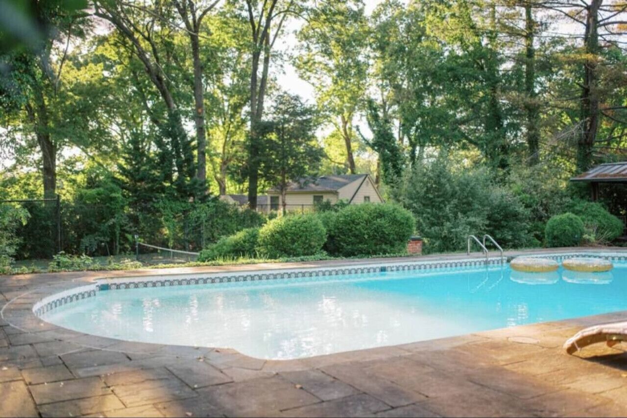 En swimmingpool med en stenbelagd poolkant, omgiven av träd och ett litet hus i bakgrunden.