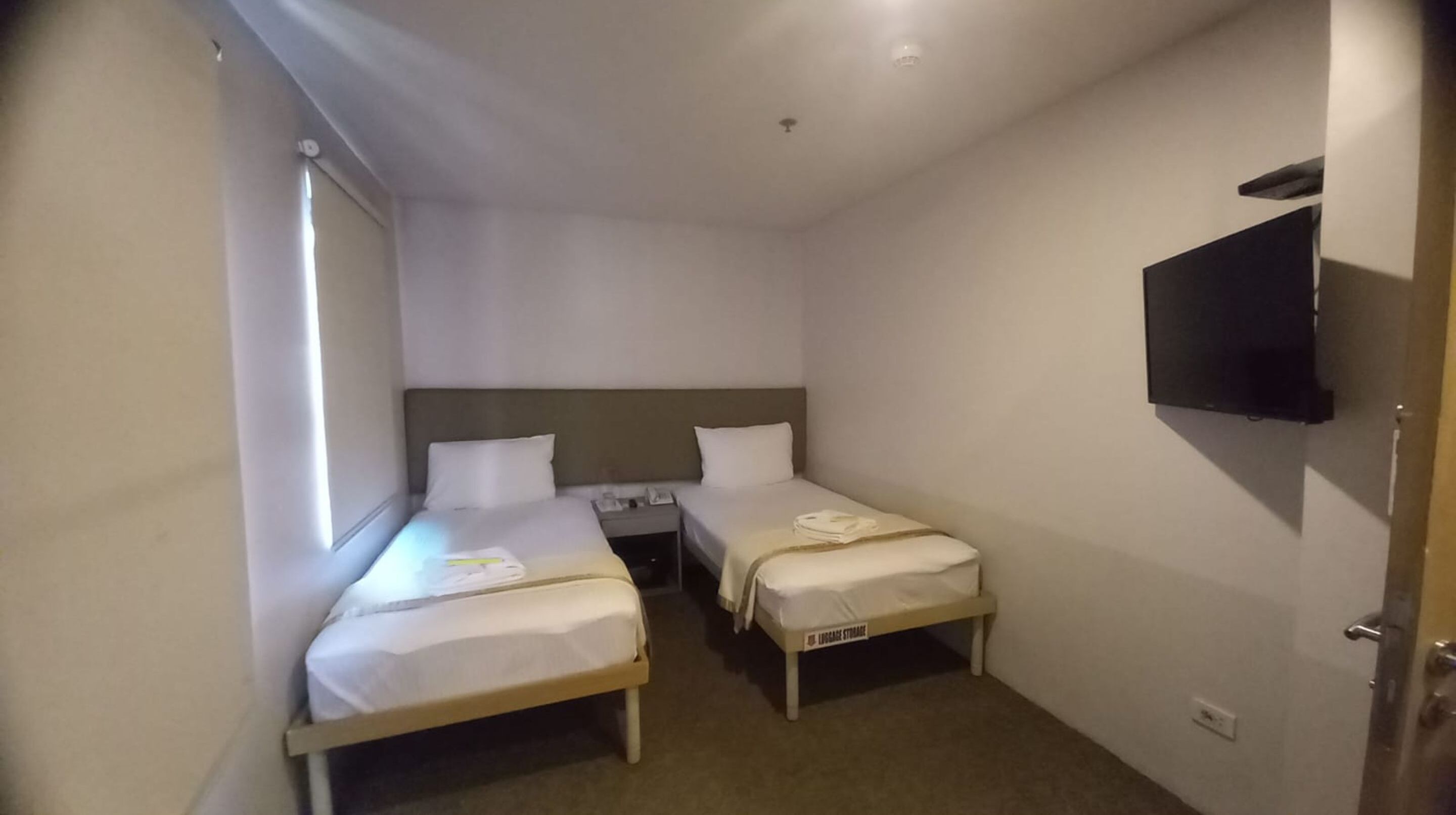 Une petite chambre d’hôtel avec deux lits simples, un téléviseur fixé au mur et une fenêtre laissant entrer la lumière naturelle.