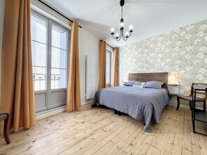 Une chambre avec un grand lit, un plancher en bois, un lustre, une fenêtre avec des rideaux et un papier peint à motifs floraux.