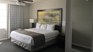 Habitación de hotel con una cama grande, ventilador de techo, un cuadro en la pared, mesita de noche con lámpara y ventana con cortinas.