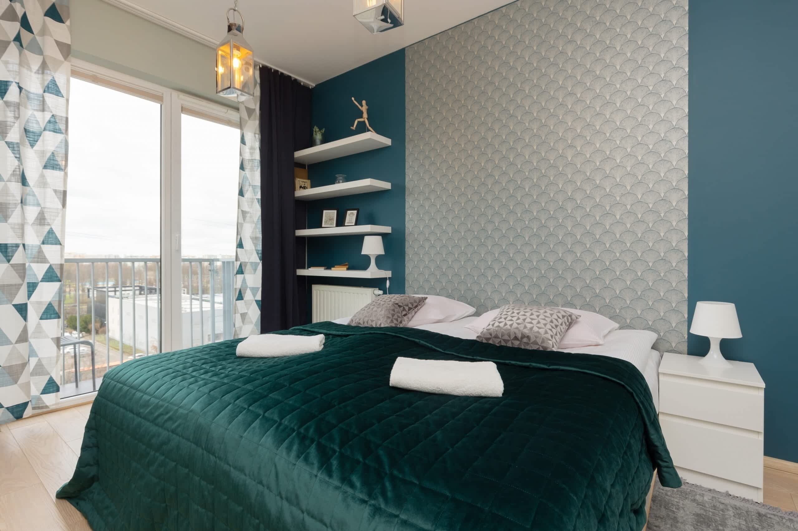 Un dormitorio moderno con una cama grande, una mesita de noche, un estante en la pared con objetos decorativos y vistas a través de una ventana.