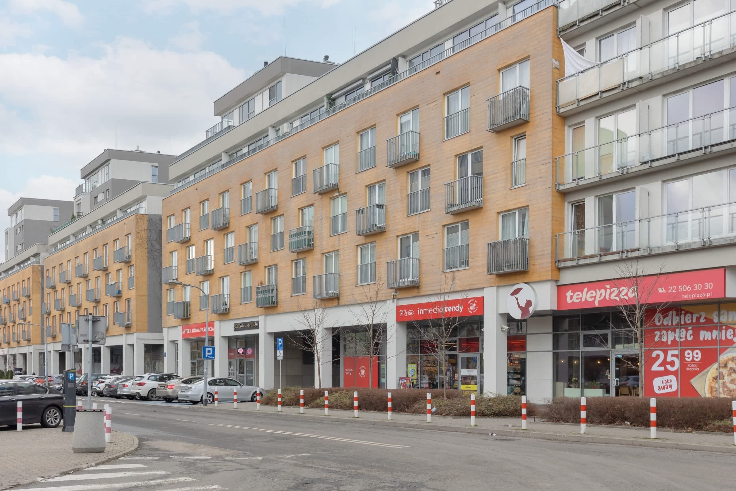 Un edificio de varios pisos con una hilera de balcones, un restaurante visible con un letrero rojo y un estacionamiento con coches.