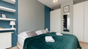 A neatly made bed with a green quilt, white pillows, and a folded towel. There is a white radiator, a gray rug, and a white shelf with a lamp. The room has teal walls and a white wardrobe. - Stylish Apartment in Wola | 450 m to the Metro, Balcony (Warszawa)