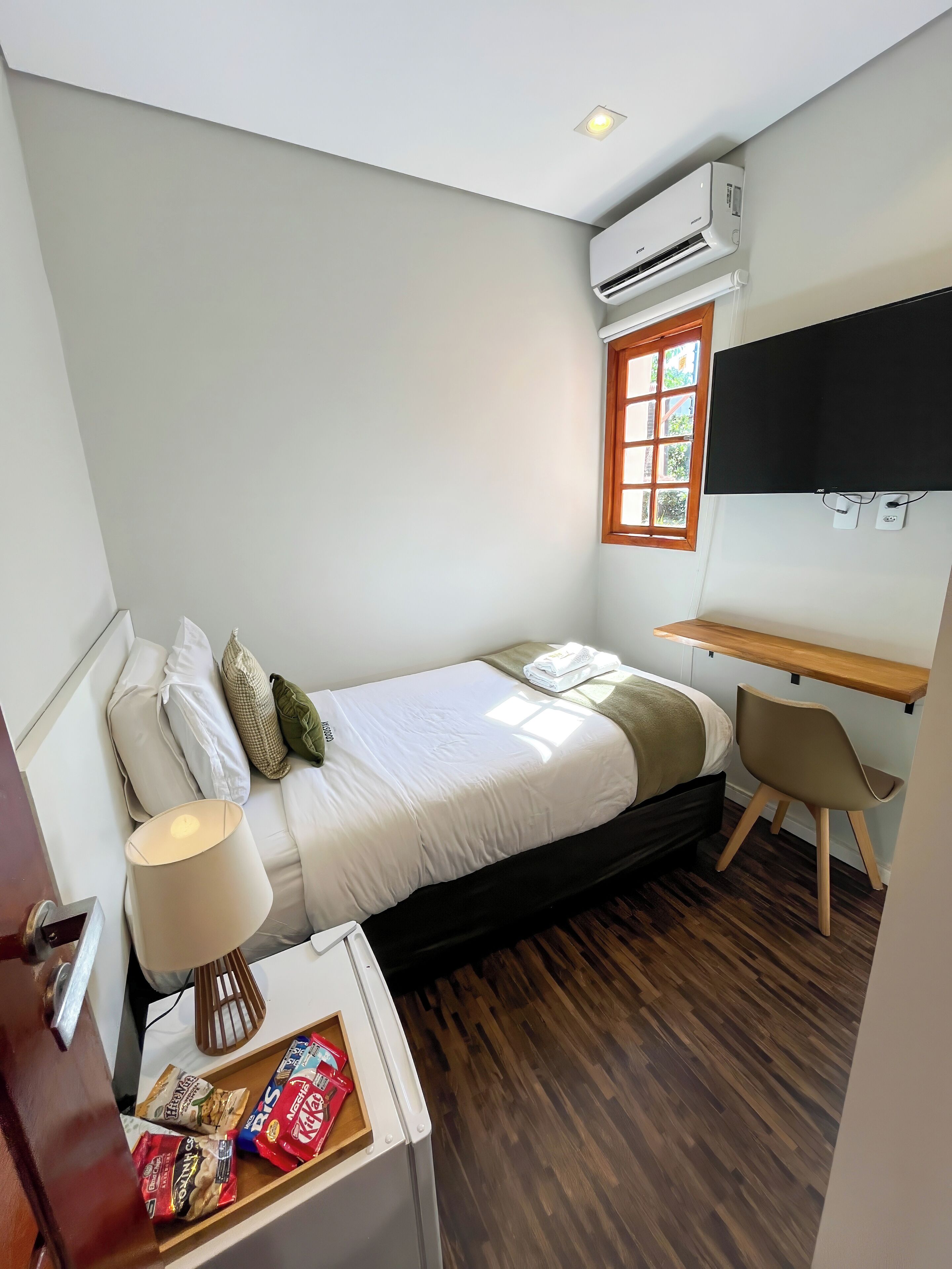 Een kleine kamer met een bed, een bureau, een stoel, een airconditioningapparaat, een televisie en een raam.