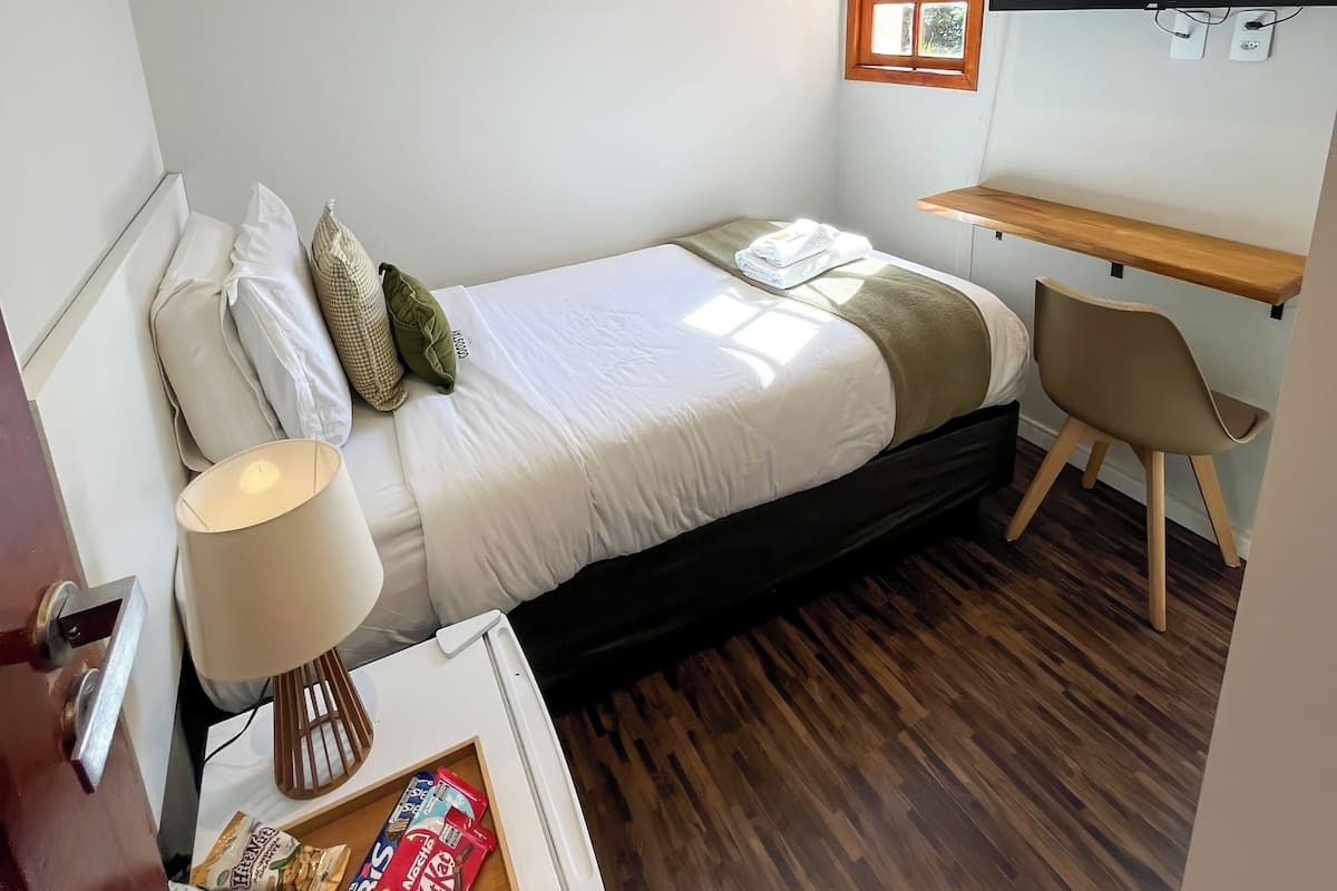 A compact room with a bed, a desk, a chair, an air conditioning unit, a TV, and a window.