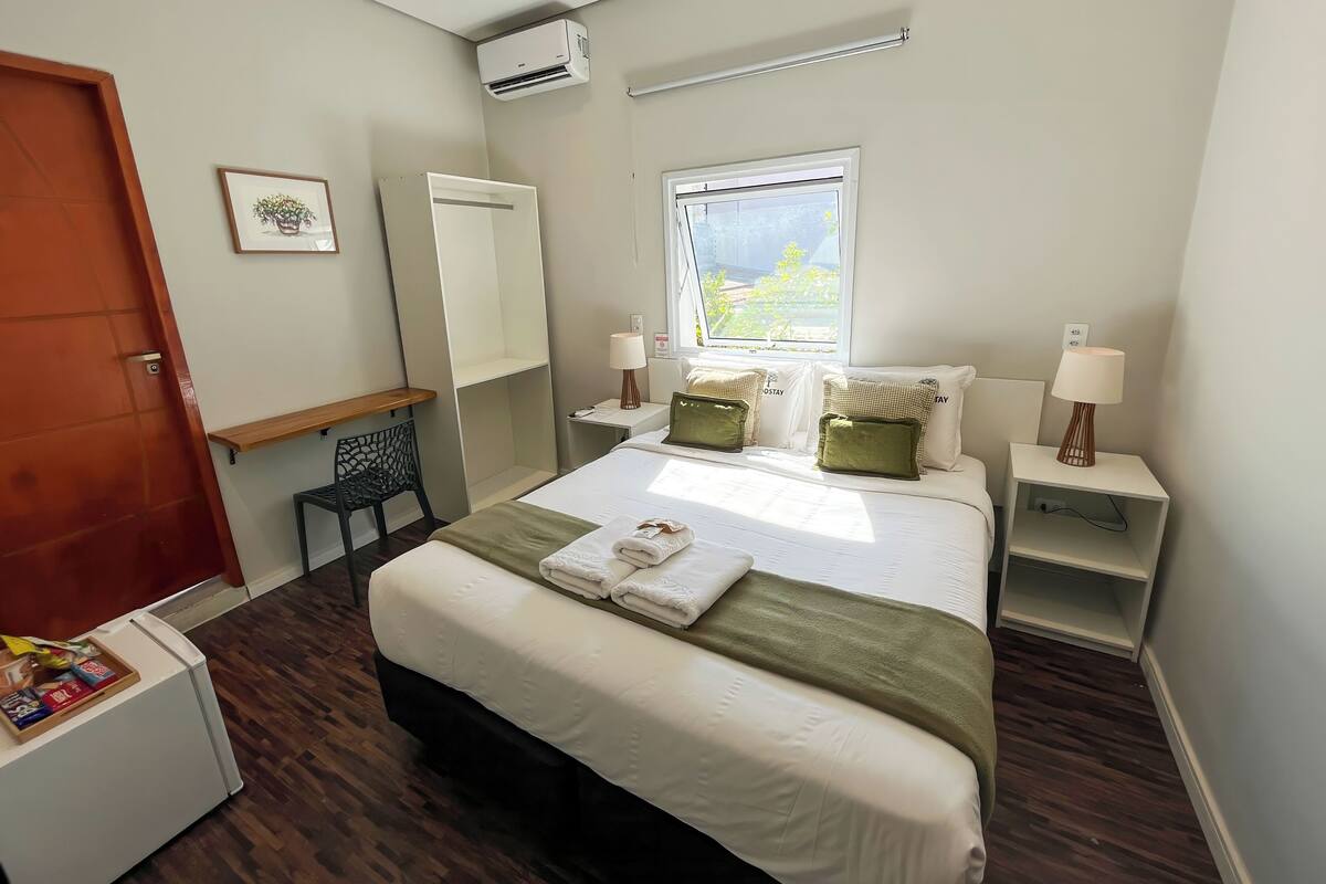 A hotel room with a bed, bedside tables, a desk, a chair, an air conditioning unit, a window, and a door.