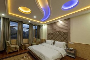 A hotel room with a large bed, a wooden headboard, a nightstand, a chair, a small table, and a view of the mountains through the windows. - Dee Grand Marq Hotel (Kargil)