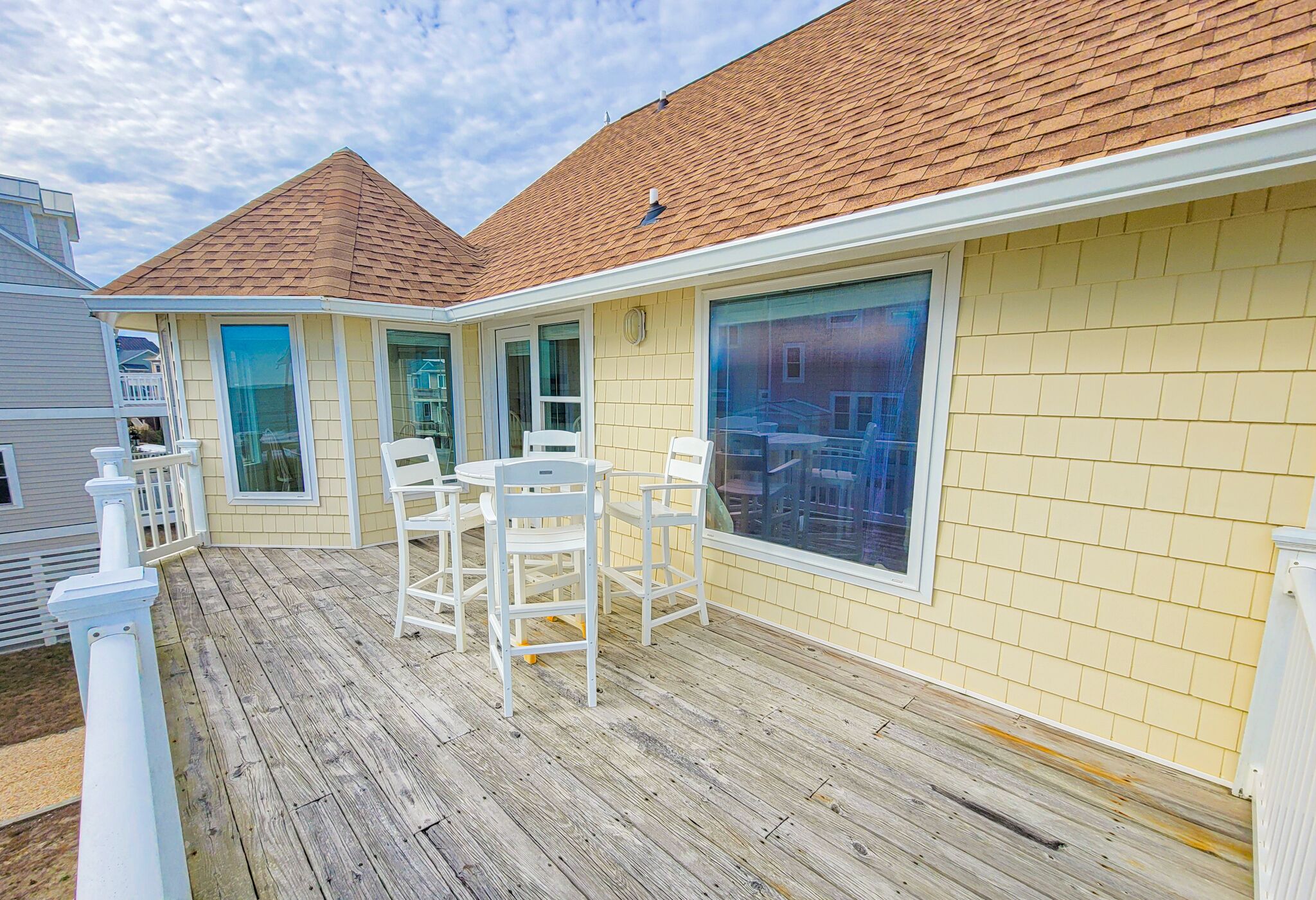 A covered outdoor deck with white chairs and a table, a yellow building with large windows, and a red-tiled roof.