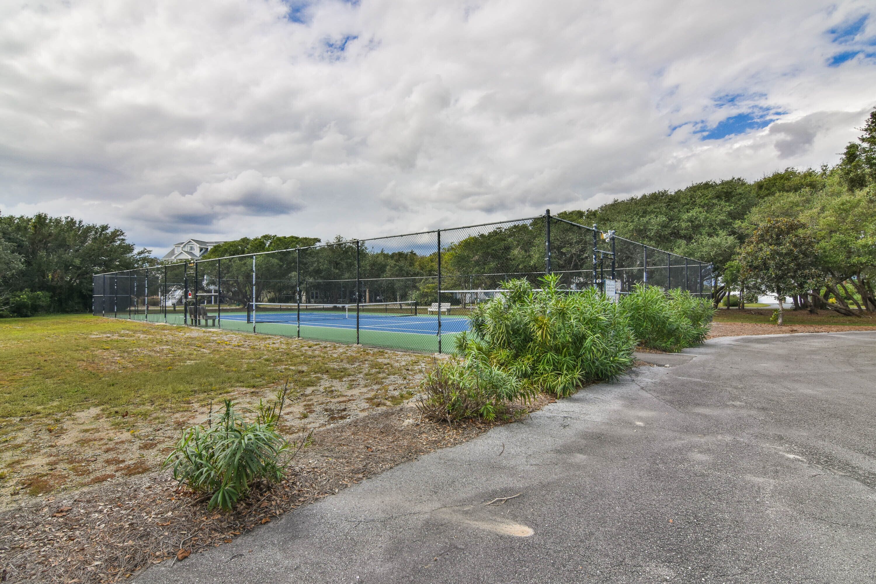Cancha de tenis con valla, rodeada de follaje y una zona pavimentada.