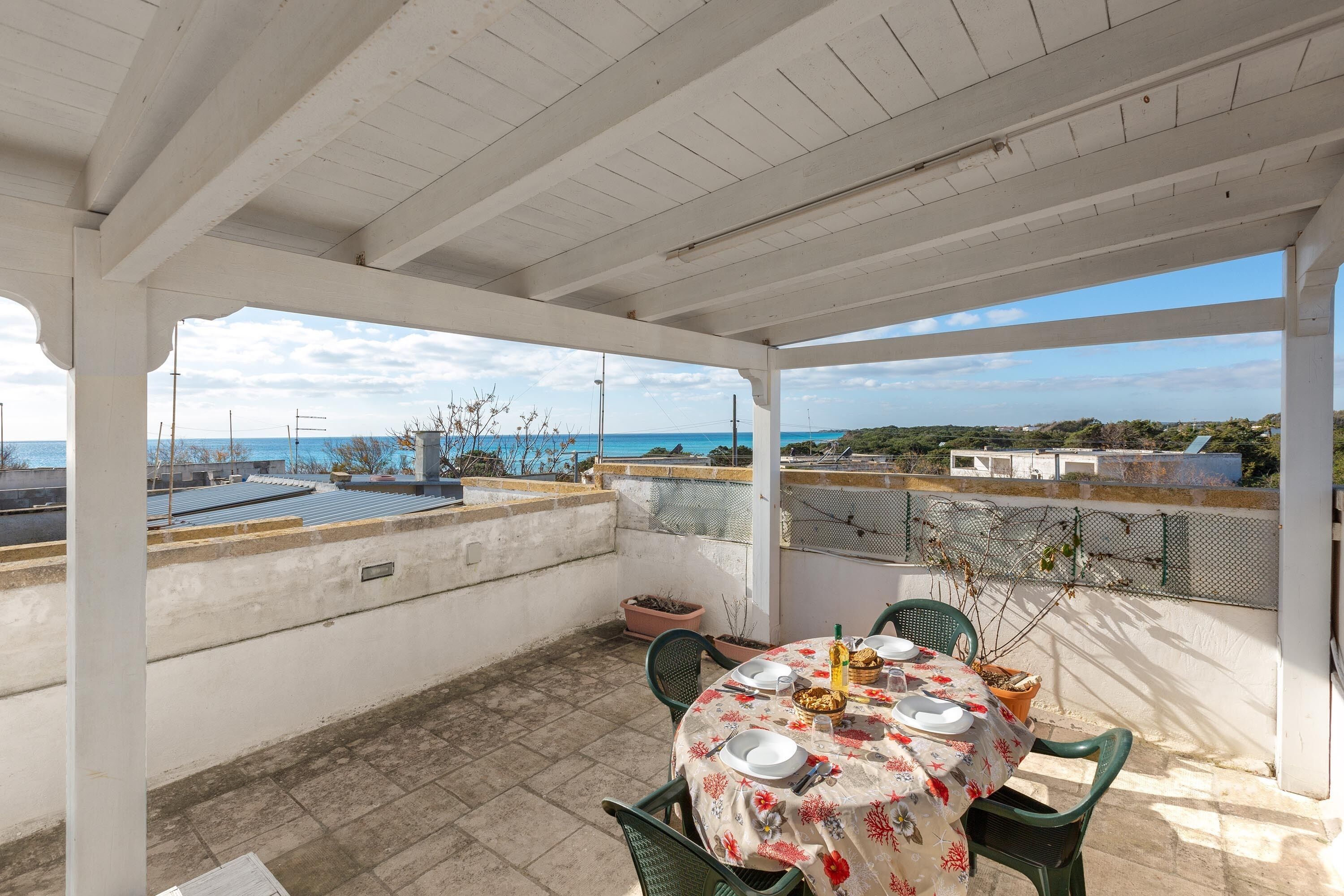 Un patio techado al aire libre con una mesa dispuesta para comer, con vistas a una zona costera con edificios y cielo despejado.