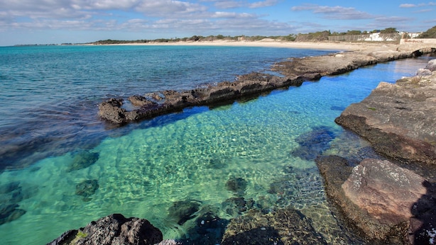 Eine Küstenansicht mit klarem türkisblauem Wasser, felsigem Ufer und einem Sandstrand.