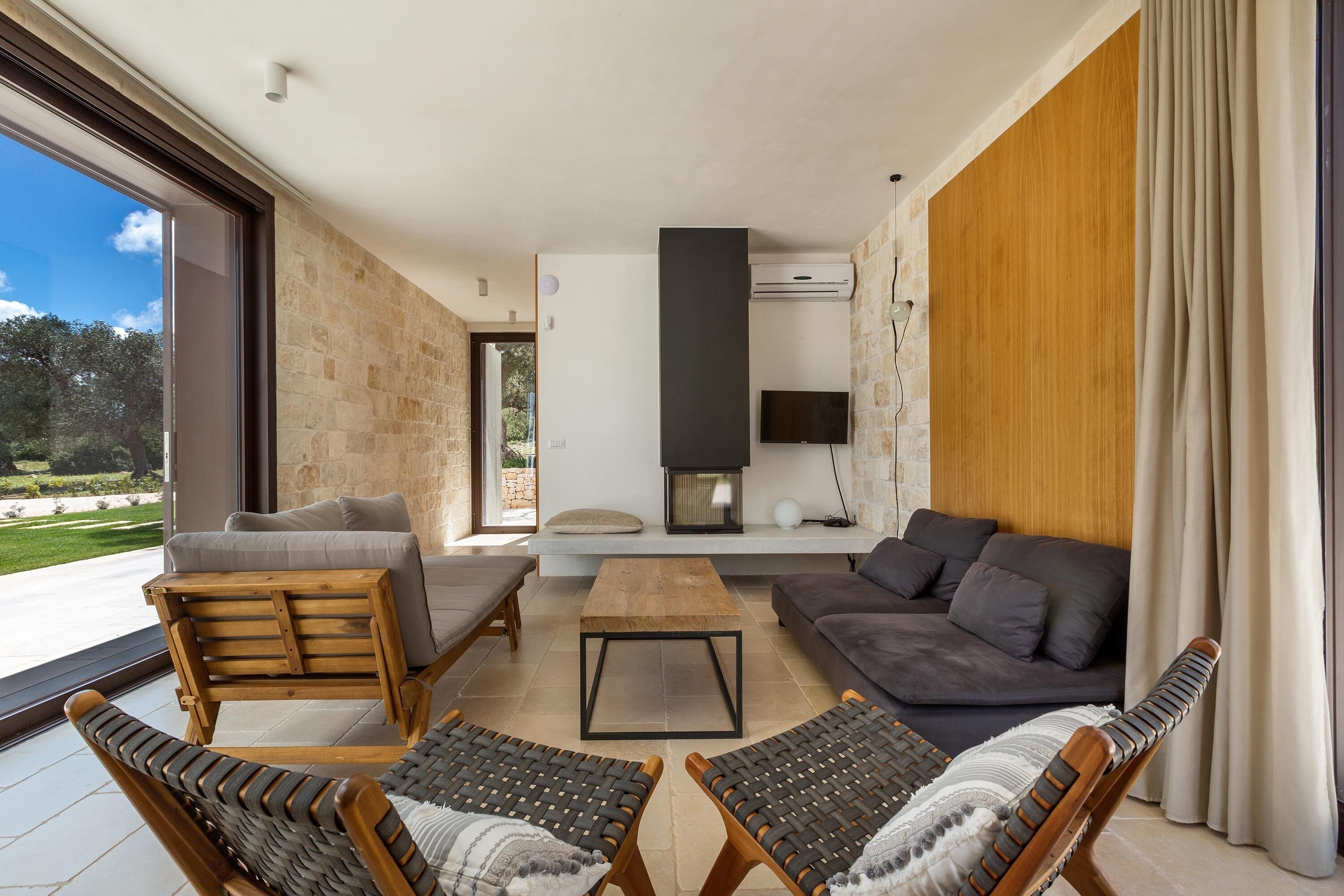 A modern living room with a stone wall, a wooden sofa, a grey couch, and two wicker chairs.