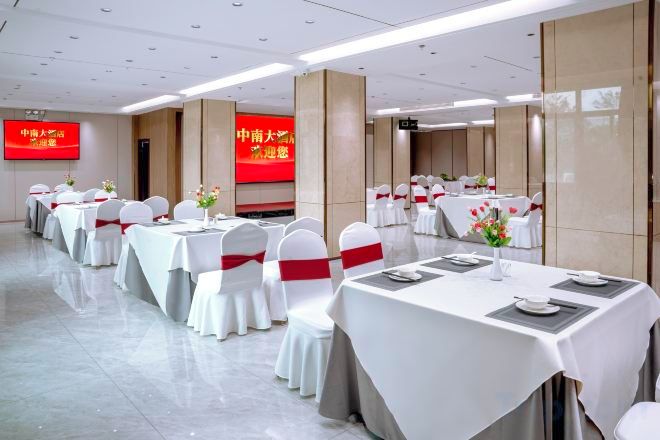 Een feestzaal met rijen tafels klaargezet voor evenementen, een groot scherm met tekst en een schoon, modern interieur.