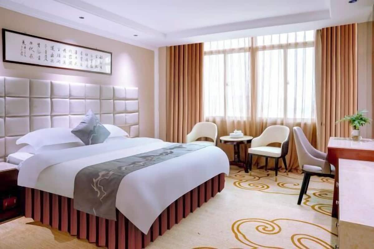 A hotel room with a large bed, a headboard with illuminated panels, a small table with chairs, and a large window with curtains.