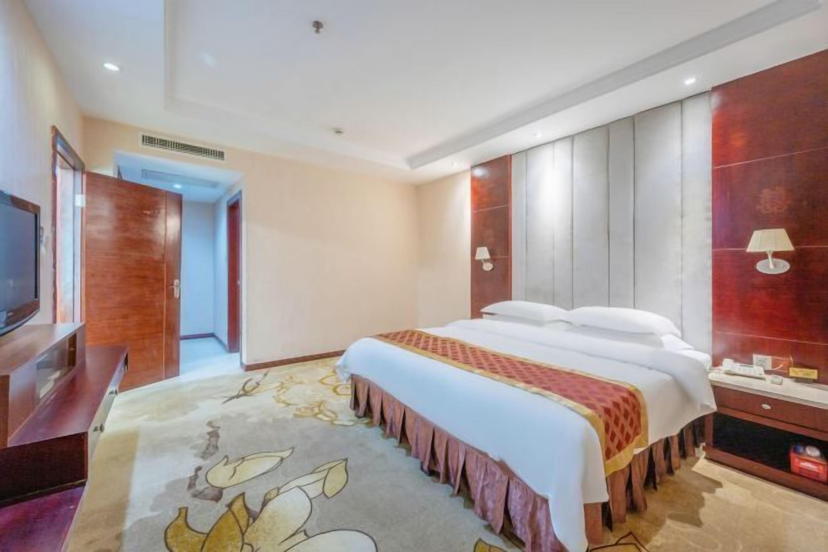 A hotel room with a large bed, a television, a wooden wardrobe, and a red accent wall.