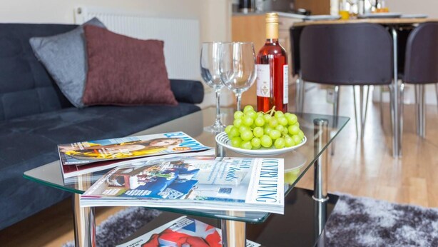 A living room with a glass coffee table holding magazines, a bottle of wine, and a bowl of grapes. - Impeccable 2-bed Apartment in Derby, England (Derby)