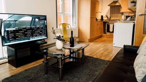 A modern living room with a flat-screen TV displaying a program guide, a dining table with a vase and wine bottle, a sofa, and a kitchen in the background. - Impeccable 2-bed Apartment in Derby, England (Derby)