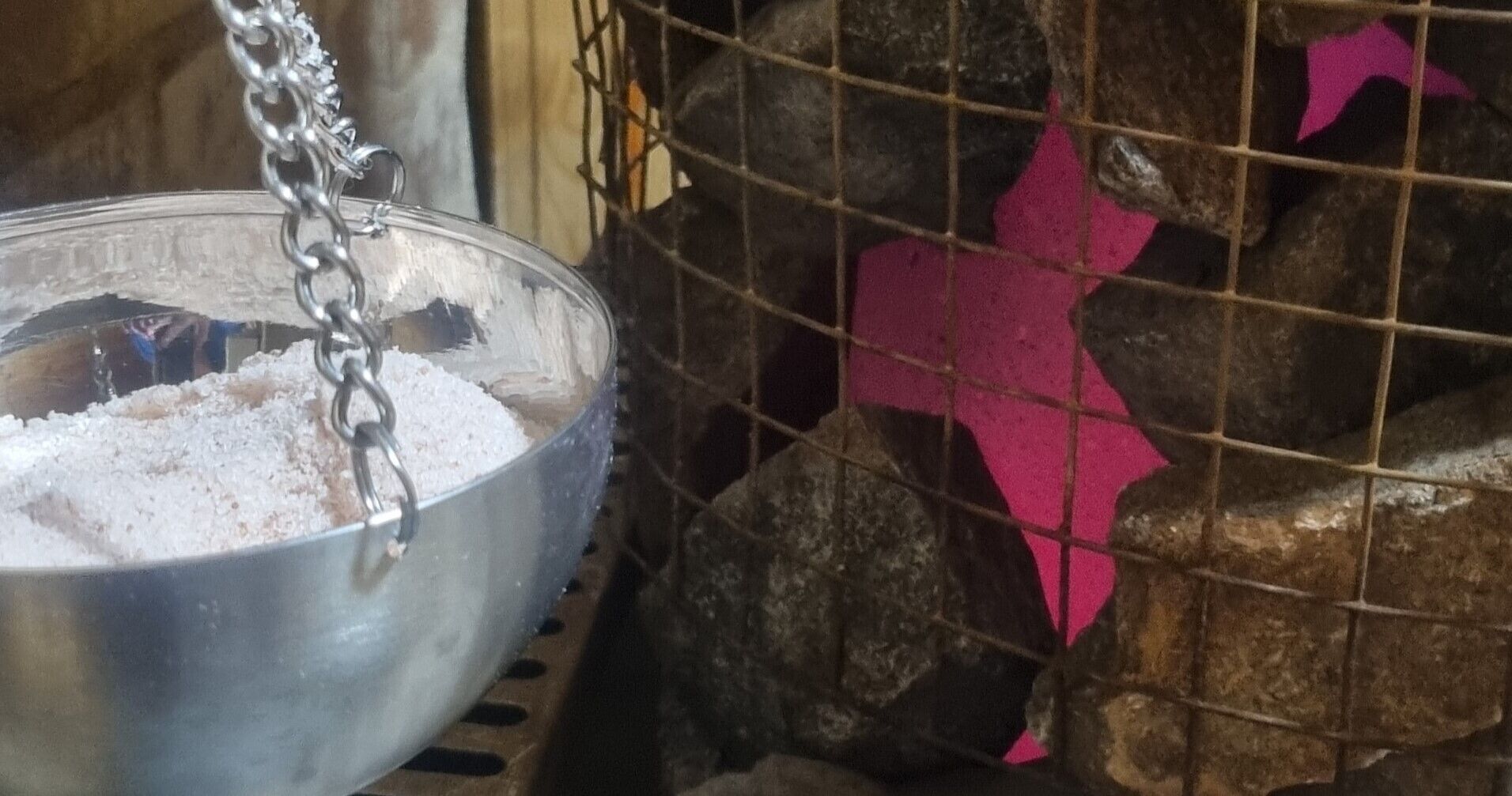 A metal bowl filled with white powder, suspended by a chain, next to a wire mesh enclosure with rocks and a pink object inside.