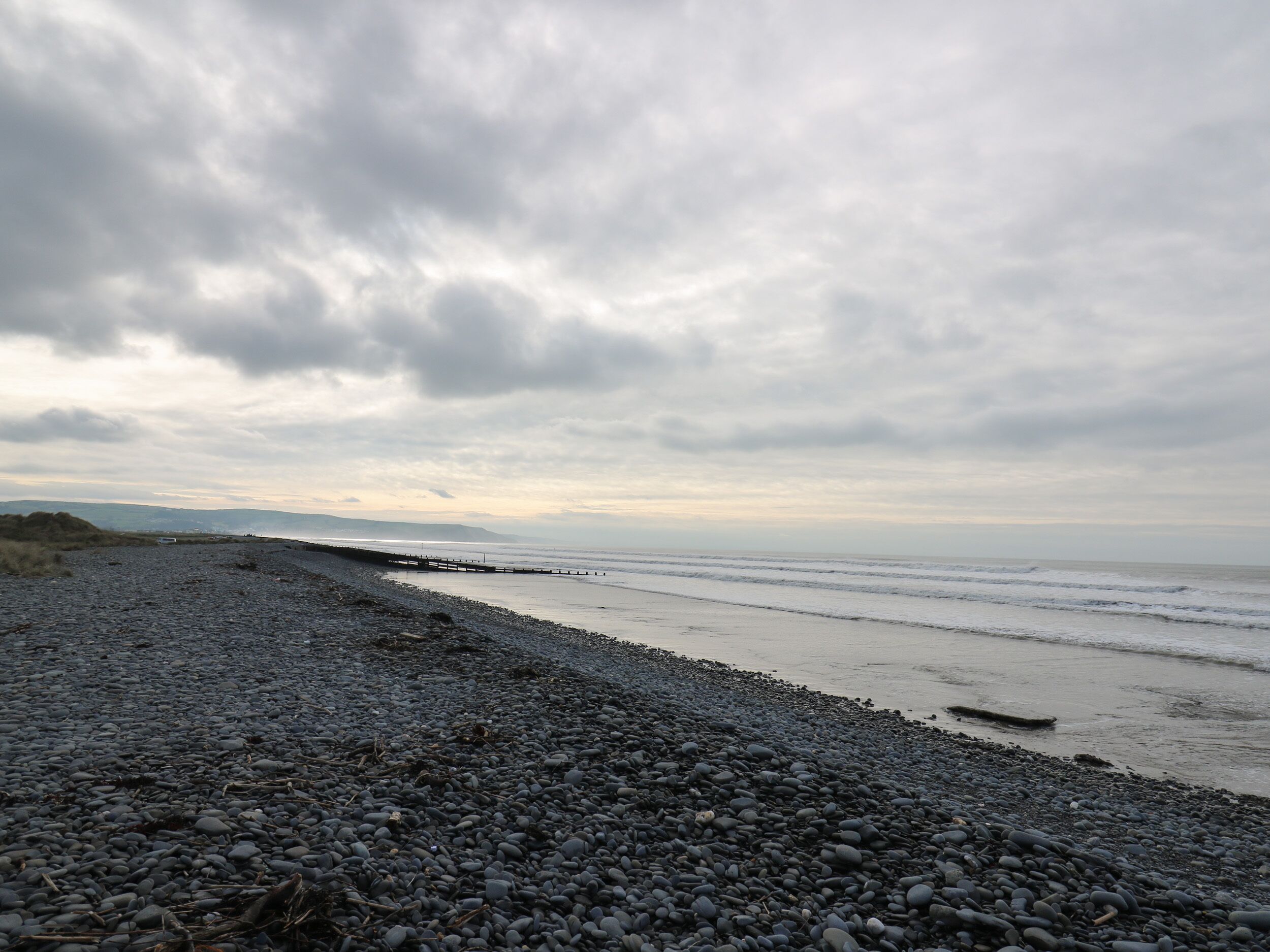 A pebbled beach with a wooden pier extending into the sea, under a cloudy sky.