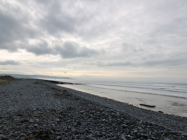 A pebbled beach with a wooden pier extending into the sea, under a cloudy sky. - Gwent 74 (Borth)