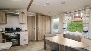 A compact kitchen with a stove, oven, and wooden cabinets. There is a dining area with a table and benches. - Gwent 74 (Borth)