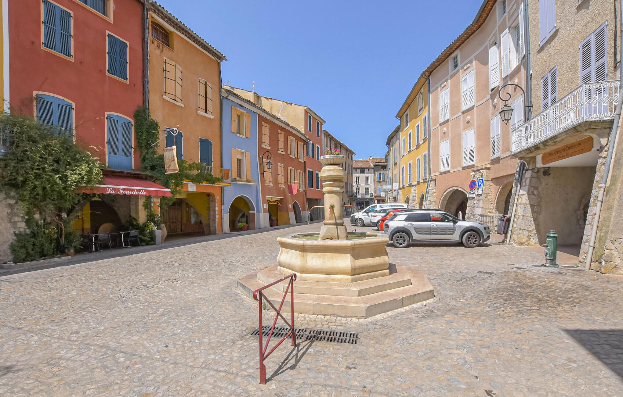 Una strada acciottolata con edifici storici, una fontana e auto parcheggiate.