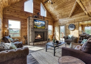 A cozy log cabin interior with a fireplace, comfortable seating, and large windows offering views of the outdoors.