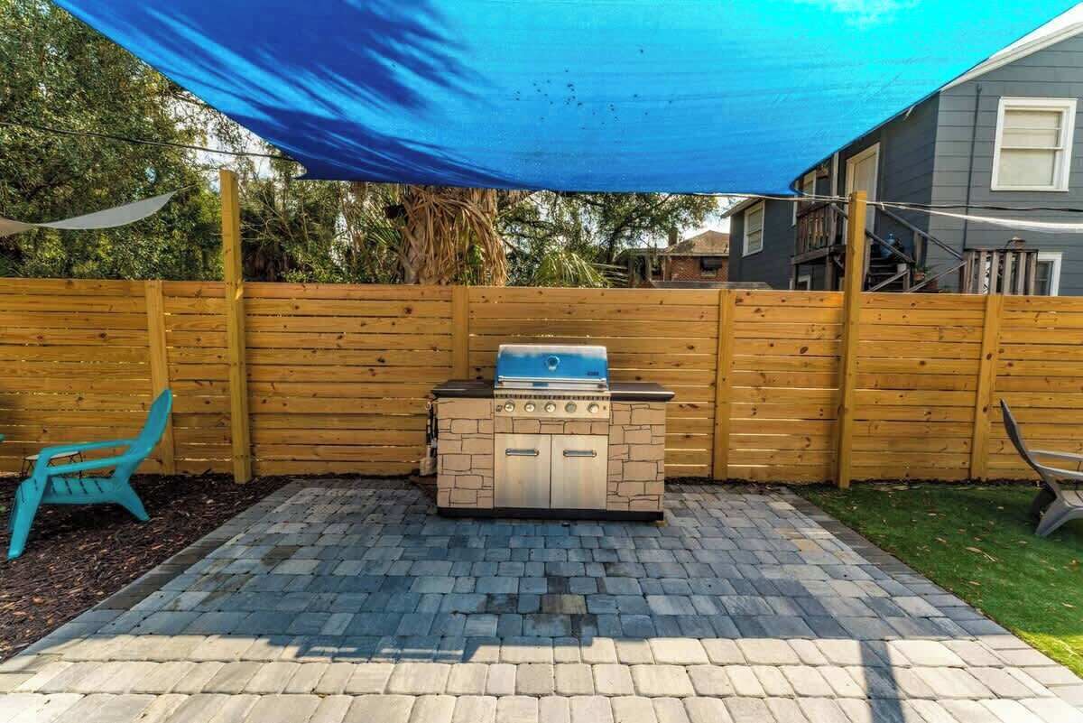Un jardin avec un barbecue, une clôture en bois et une voile d’ombrage bleue.