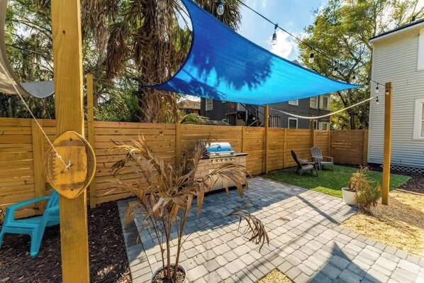 A backyard with a wooden fence, a barbecue grill, a blue shade sail, and a potted plant.