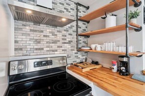 A kitchen with a modern stove, a tiled backsplash, wooden shelves, and a coffee maker.