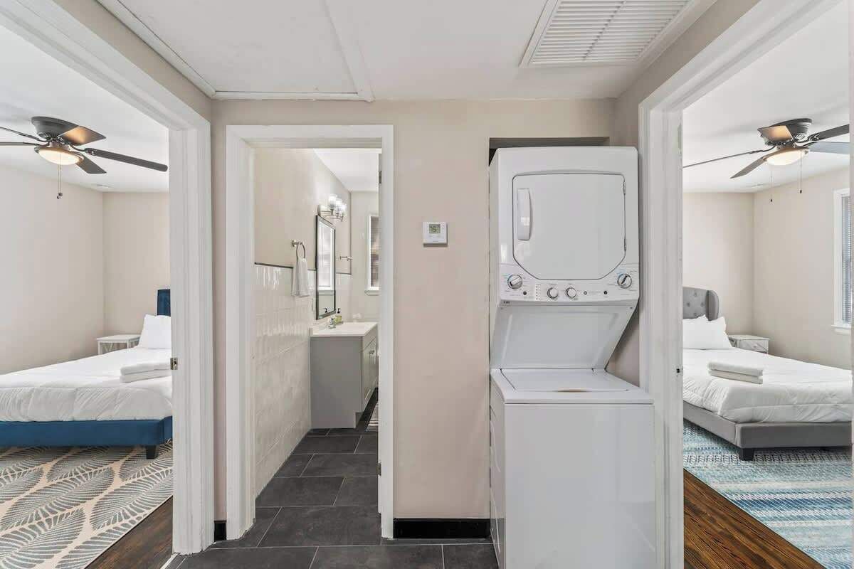 A hotel room with a bed, a washing machine, a ceiling fan, and a bathroom with a mirror and sink.