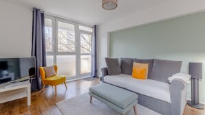 Un salon moderne avec un canapé gris, un fauteuil jaune, un téléviseur posé sur un meuble blanc et une grande fenêtre avec des rideaux.
