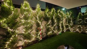 Zona ajardinada con árboles de Navidad artificiales decorados con luces, un muro de piedra y una pequeña mesa redonda con una planta en maceta.