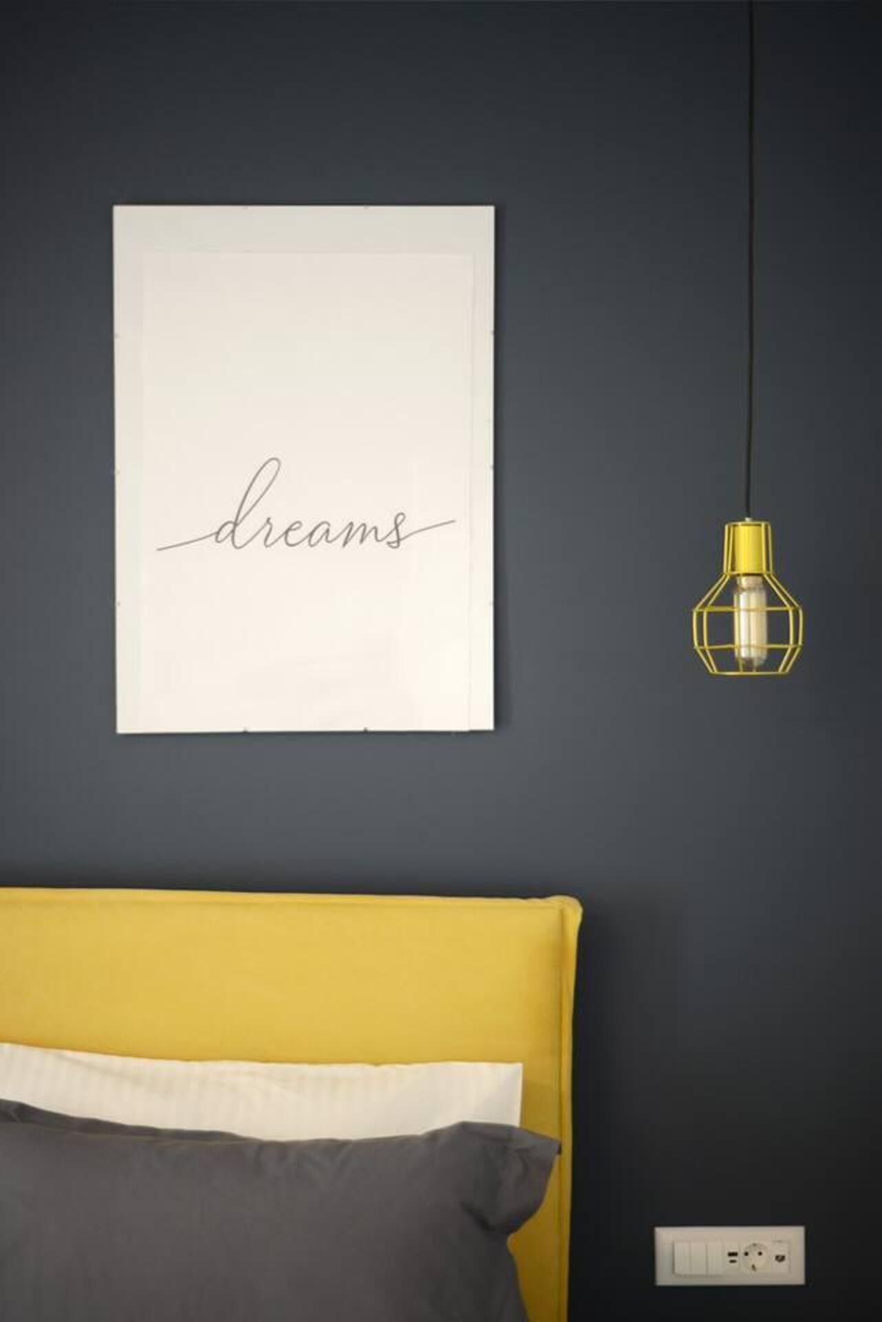 A room with a yellow headboard, a gray pillow, a framed word "dreams" on the wall, and a hanging yellow light fixture.