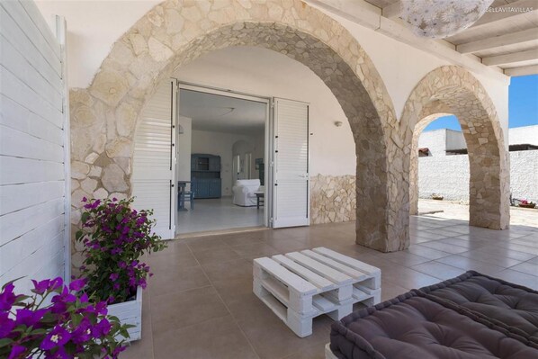 A covered outdoor area with stone arches, a white door leading to a room, and a wooden bench.