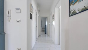 Un couloir aux murs blancs, doté d’un sol carrelé et d’une porte au fond qui donne sur un espace cuisine.
