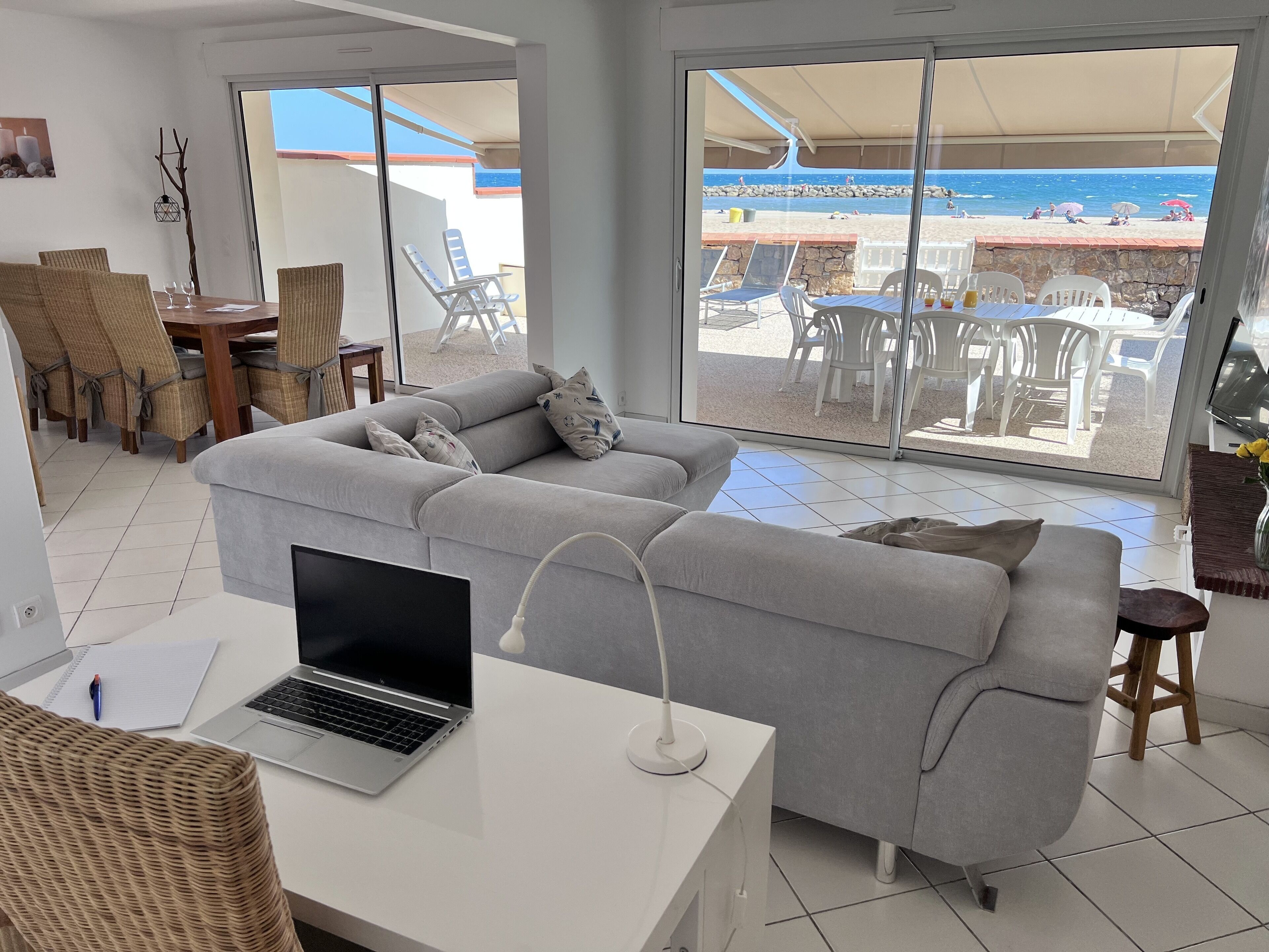 Een moderne woonkamer met een grijze bank, een laptop en een eethoek met rieten stoelen.