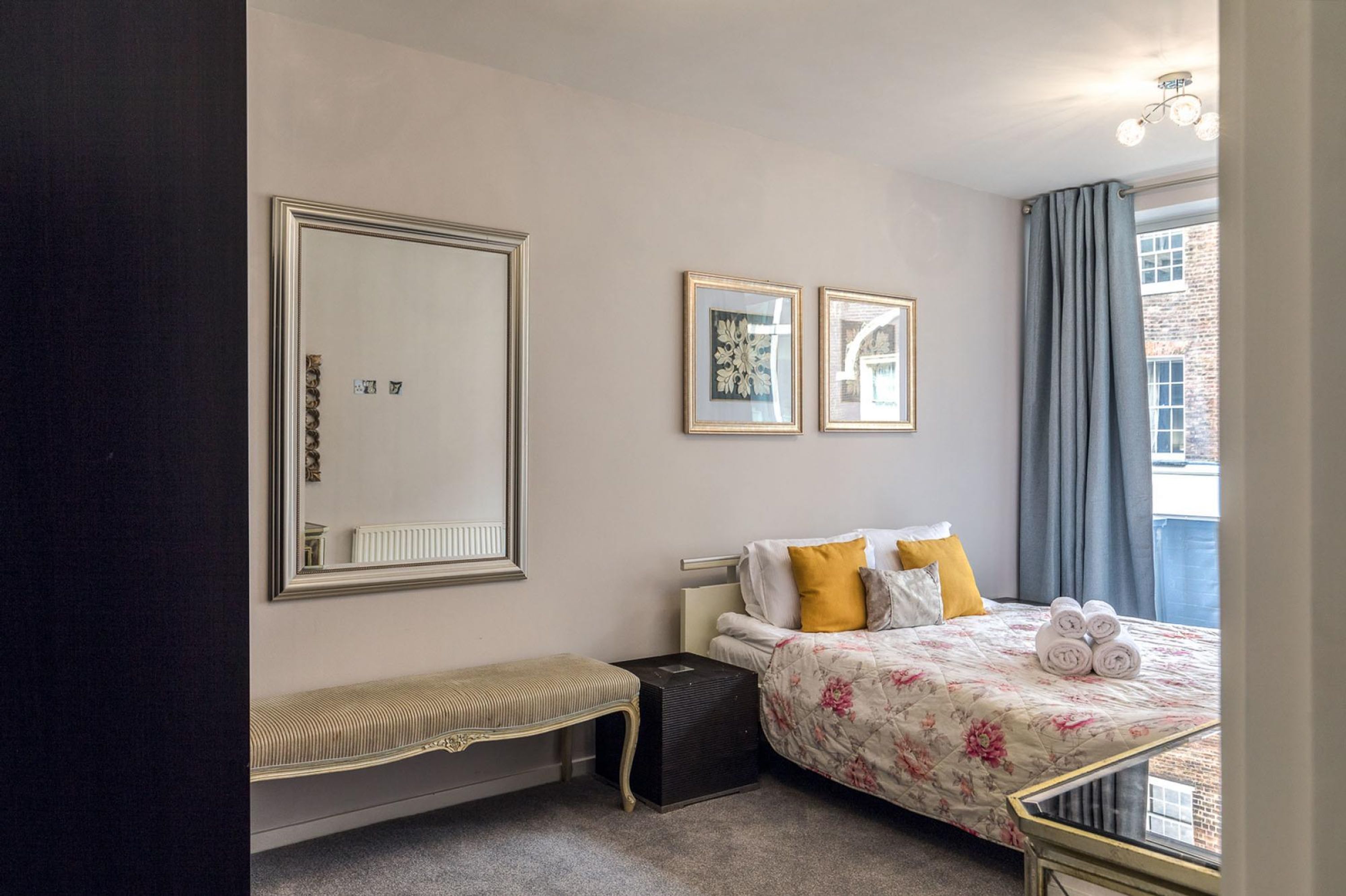 A hotel room with a large mirror, two framed pictures, a bed with floral bedding, a bench, and a radiator.